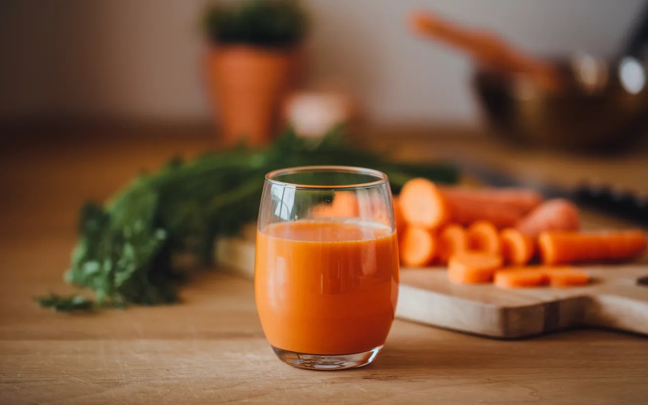 A glass of fresh carrot juice sits on a wooden table with chopped carrots and carrot greens in the background.