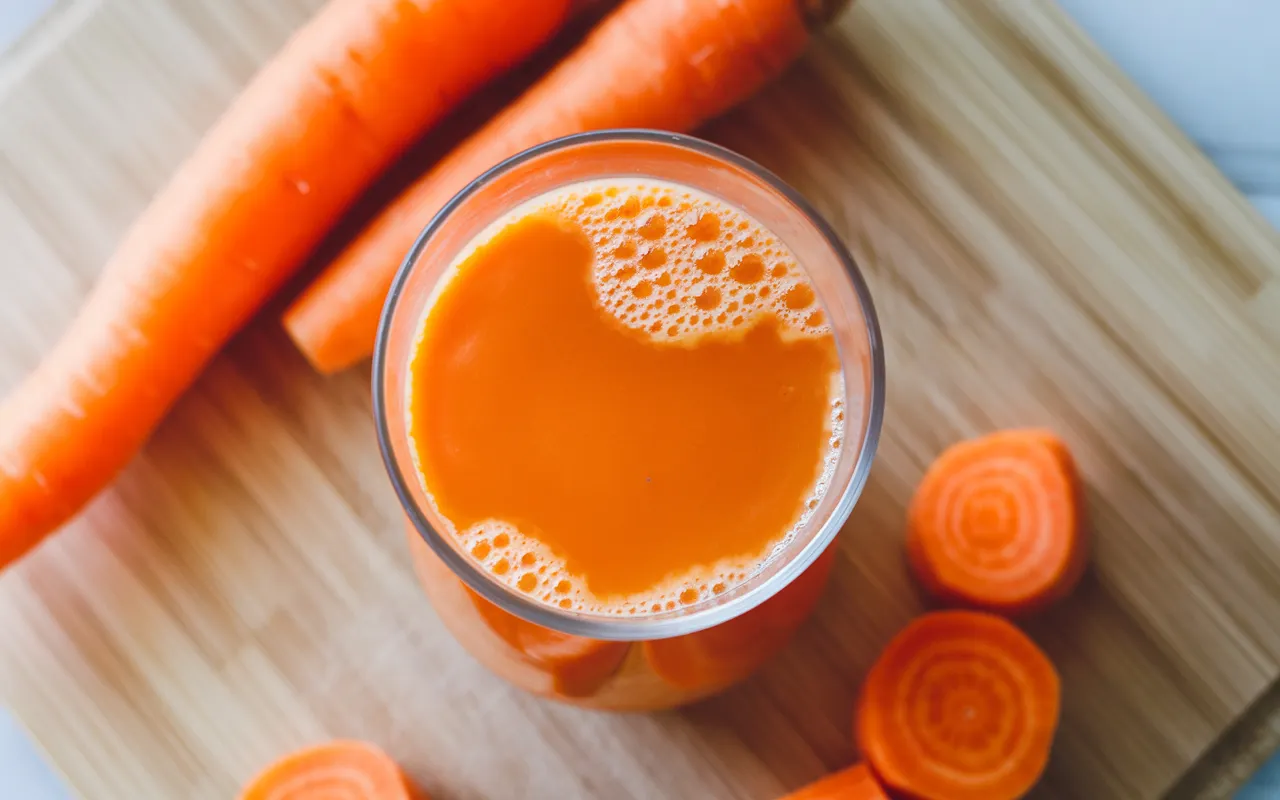 A glass of fresh carrot juice next to three whole carrots on a wooden table, showcasing the natural ingredients needed for the beverage.